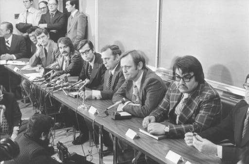 Suited men sit along a long table