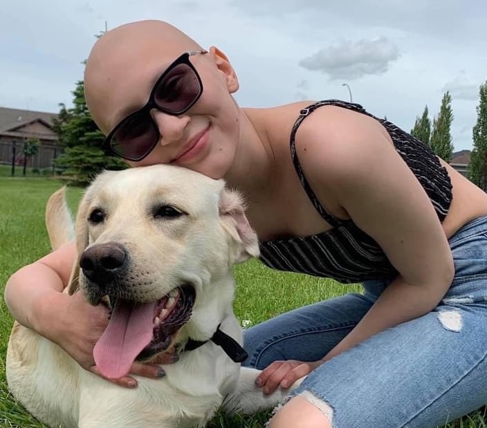 A teenager wearing jeans, a tank top and sunglasses, is kneeling on grass, posing with her dog.